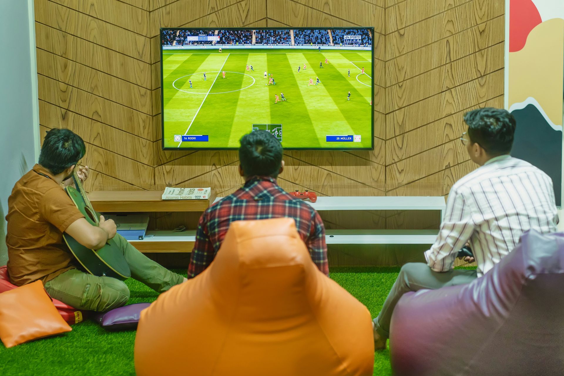 Three young men watch a soccer video game on TV in a game room, one playing guitar.