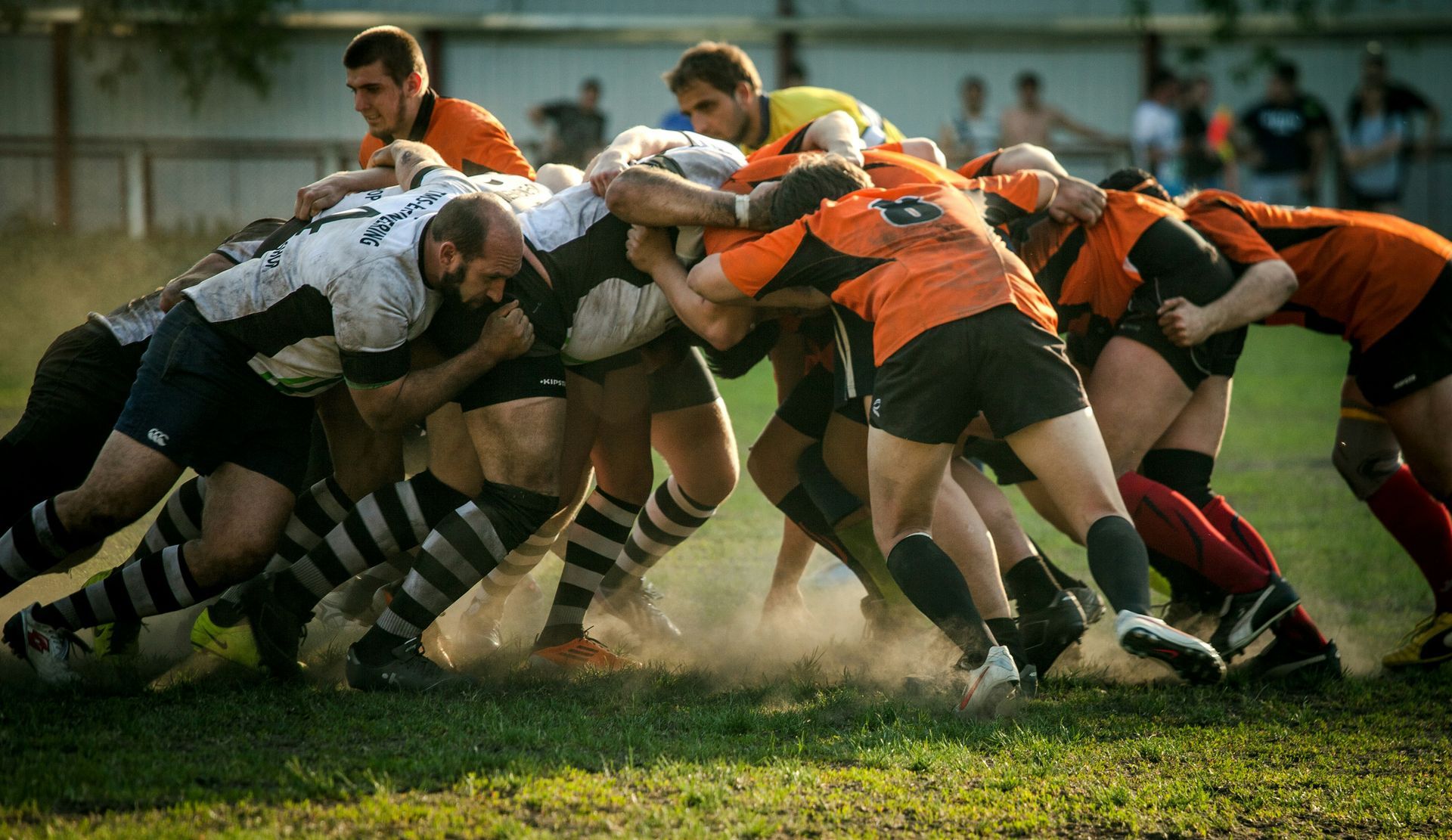 Rugby players in a scrum, engaged in a tight, physical contest on a grass field. Players wear black and orange jerseys.