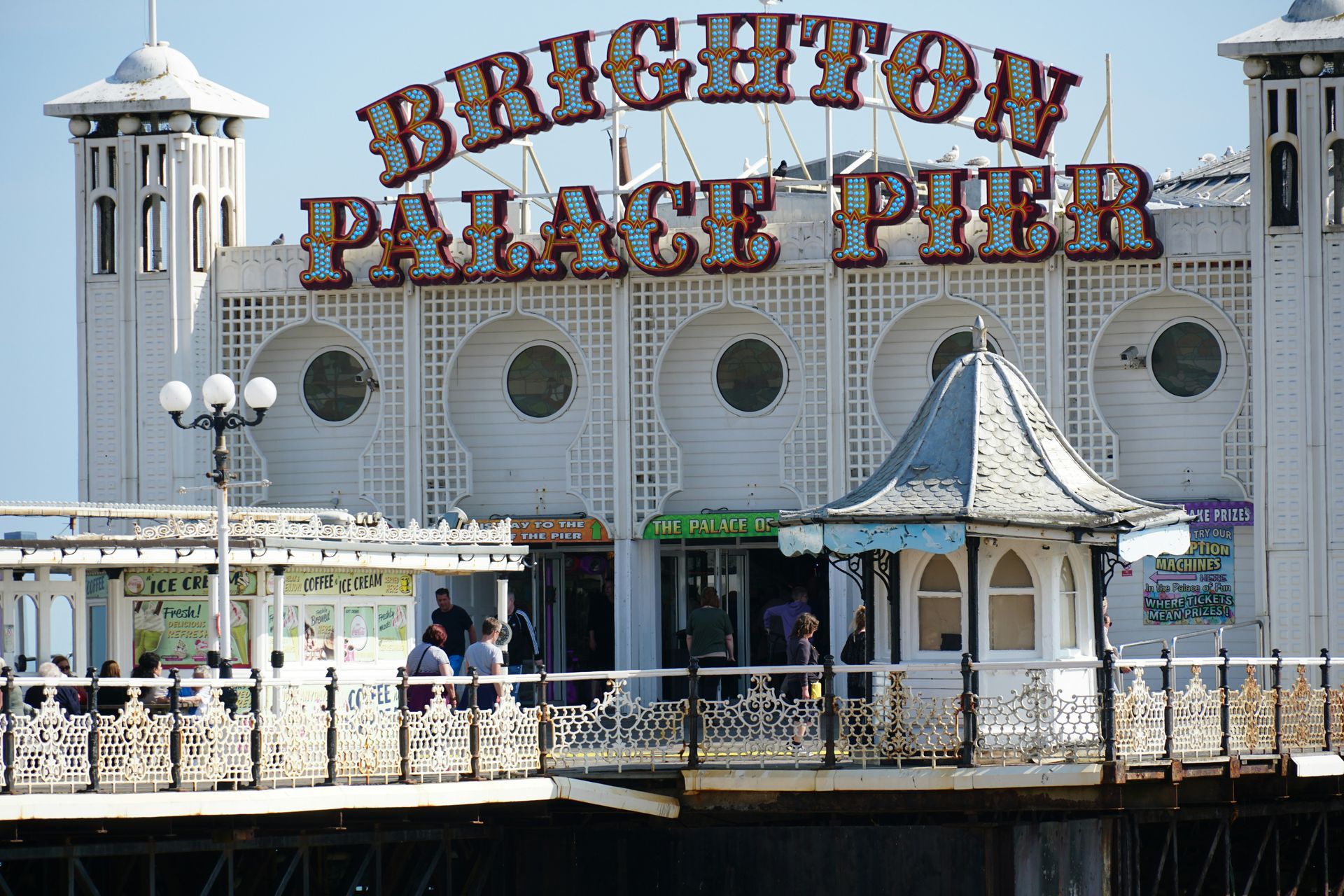 Brighton Palace Pier entrance, white building with sign, people near entrance, blue sky.