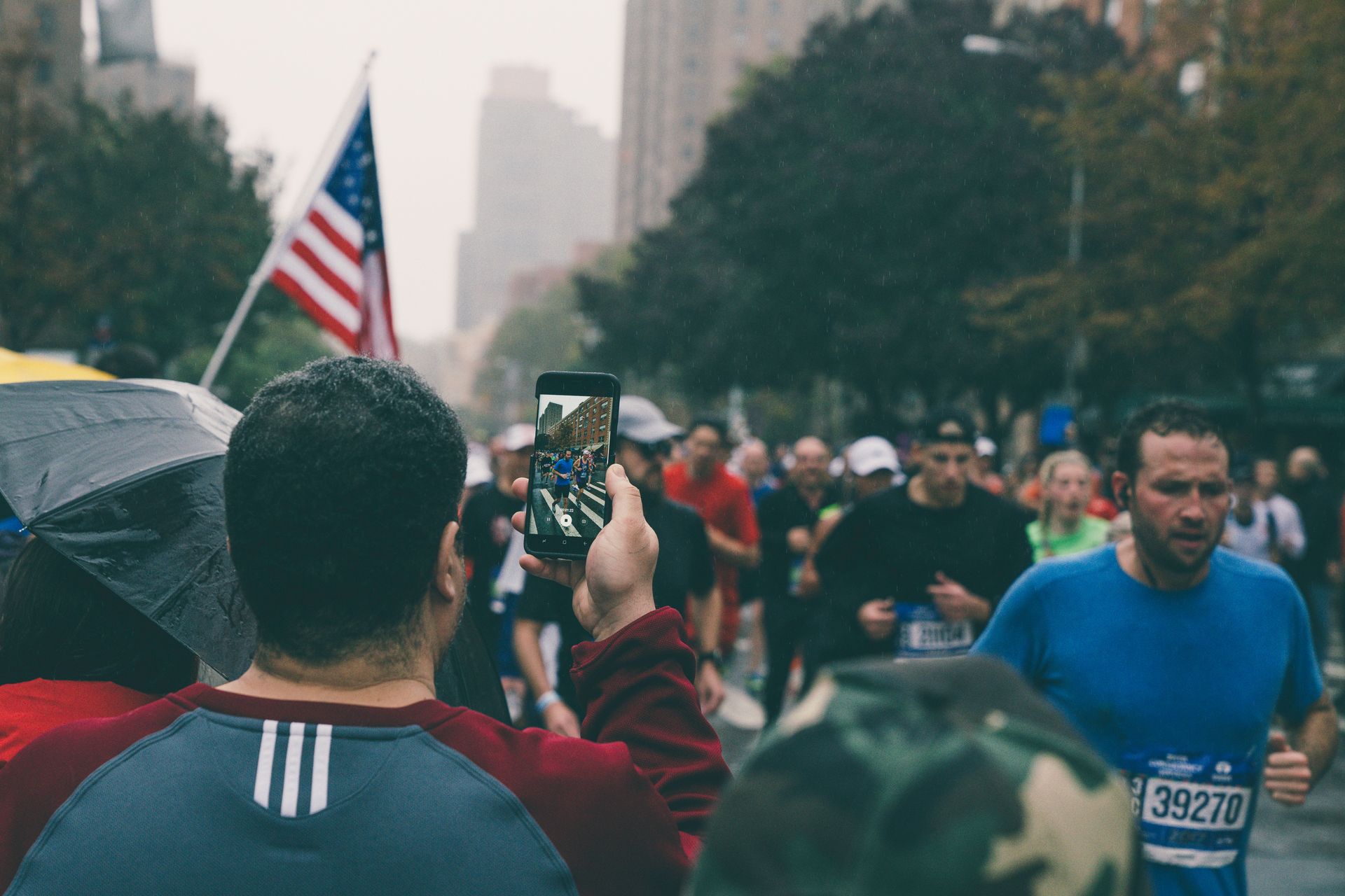 Spectator films marathon runners with a phone; American flag and cityscape in the background.