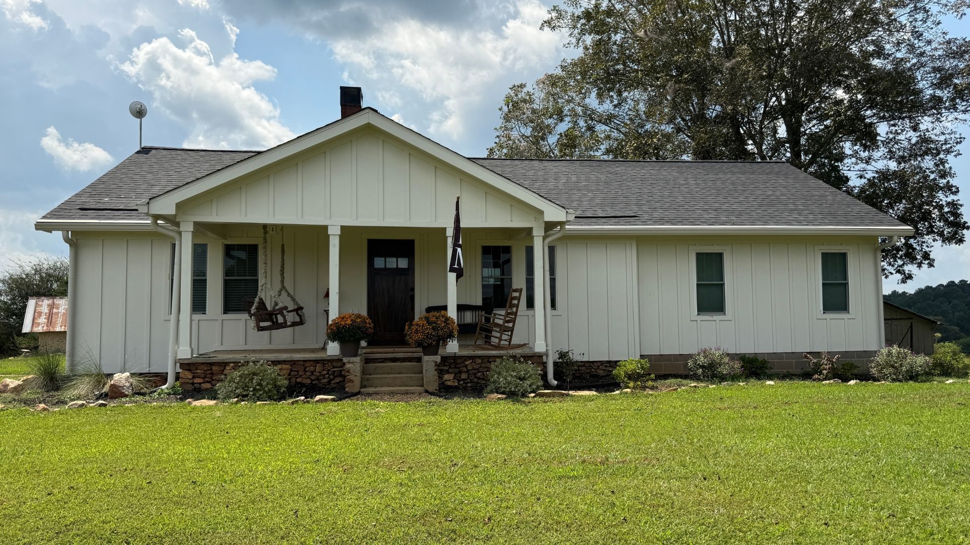 White farmhouse with porch, black roof, set in a grassy field under a cloudy sky.