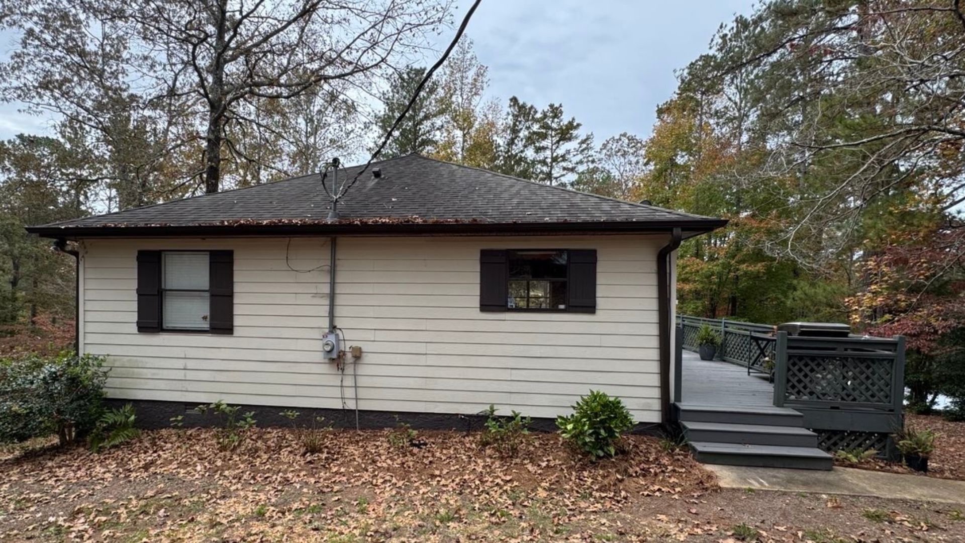 Small, beige house with dark brown trim, shutters, and a deck. Overcast sky, surrounded by trees and leaves.