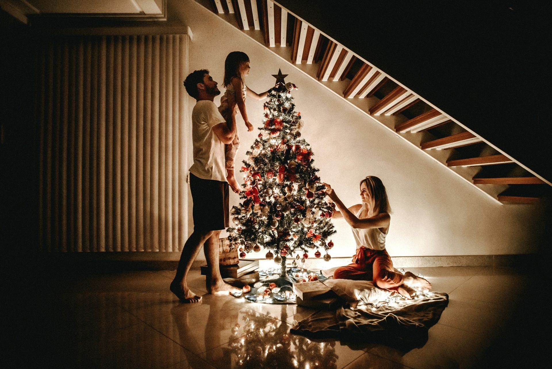 Family decorating Christmas tree: father holding child, mother kneeling, stairs in background.