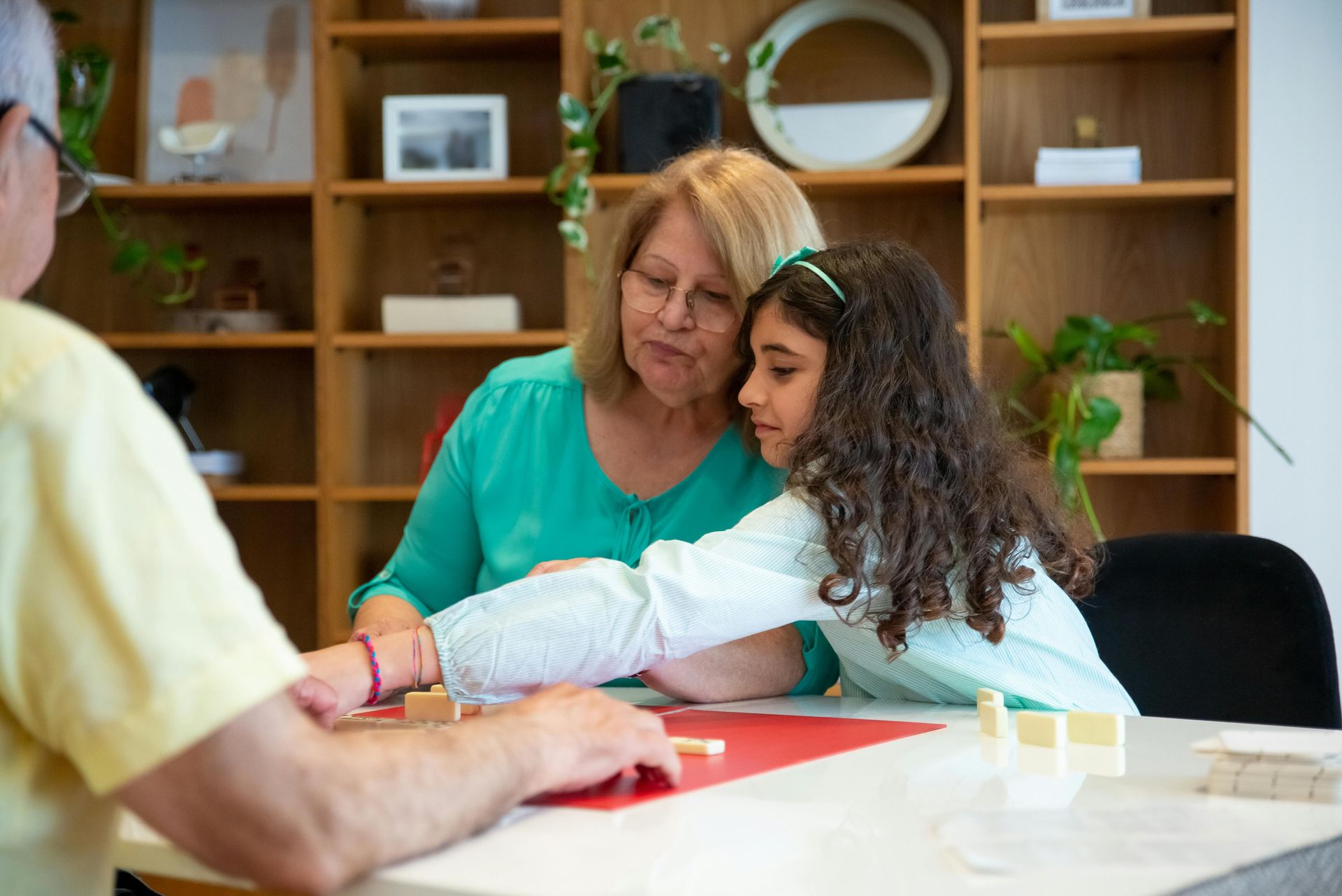 Grandmother and girl playing a game at a table with an older man, in a room with shelves.