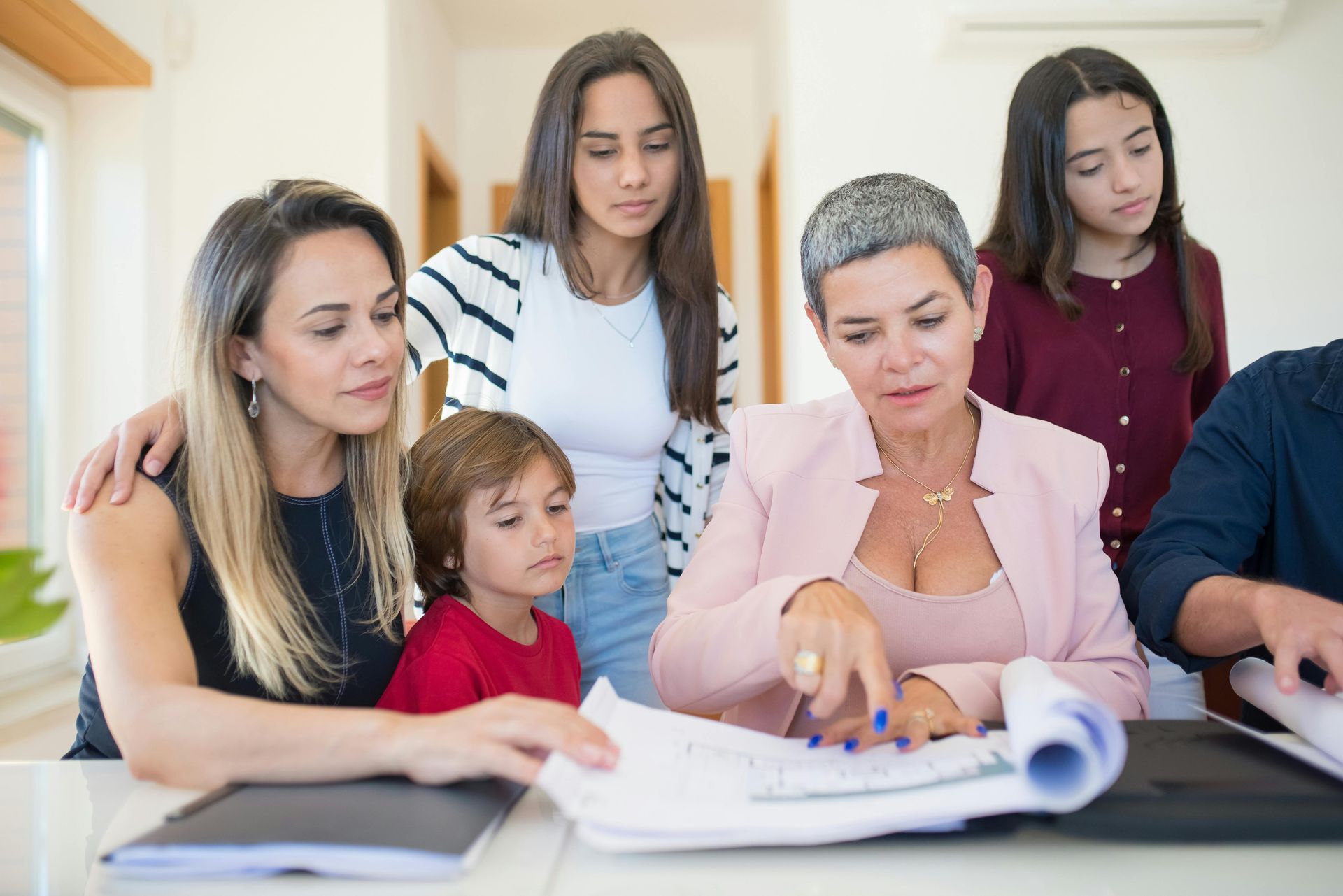 A group gathers around a table, looking at documents while a person in a pink blazer points to the paperwork.