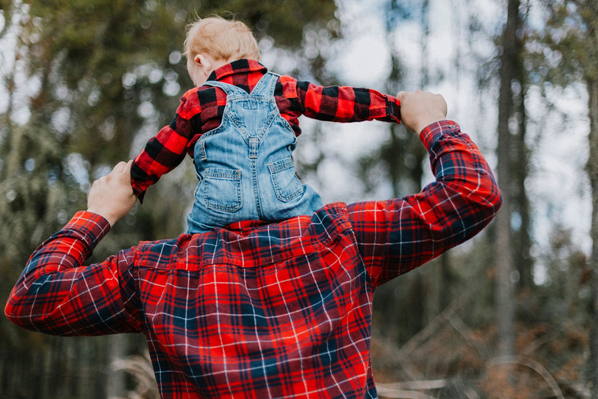 A person carries a child on their shoulders in a forest setting; both are wearing red plaid.