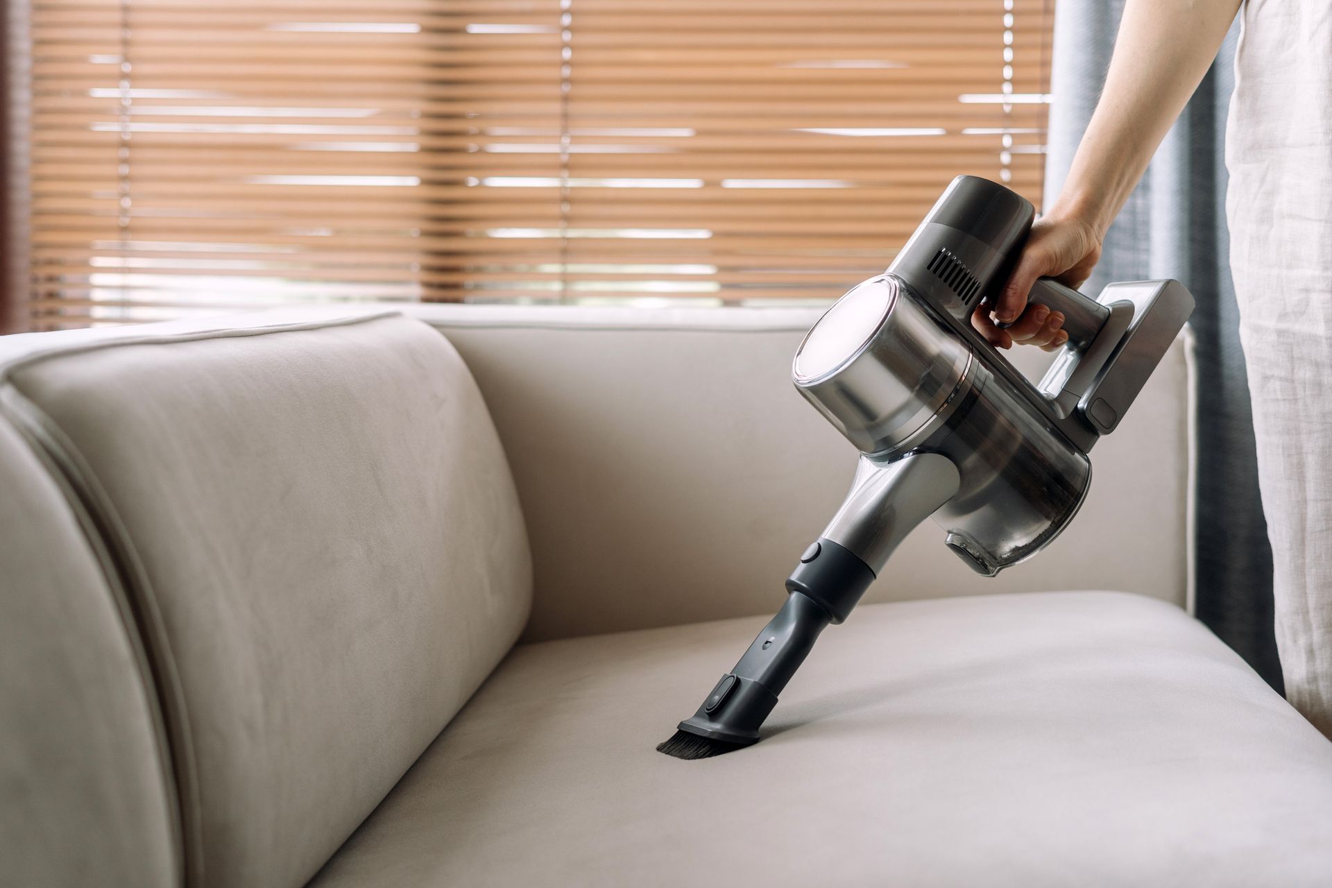 Close-up of a hand operating a handheld vacuum to clean a light-colored sofa.