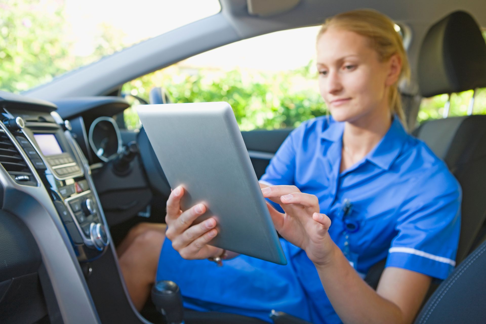 A nurse is sitting in the driver 's seat of a car using a tablet.