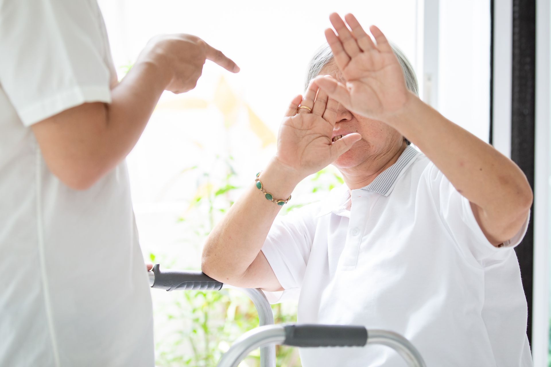 An elderly woman is sitting in a wheelchair with a walker and a nurse is pointing at her.