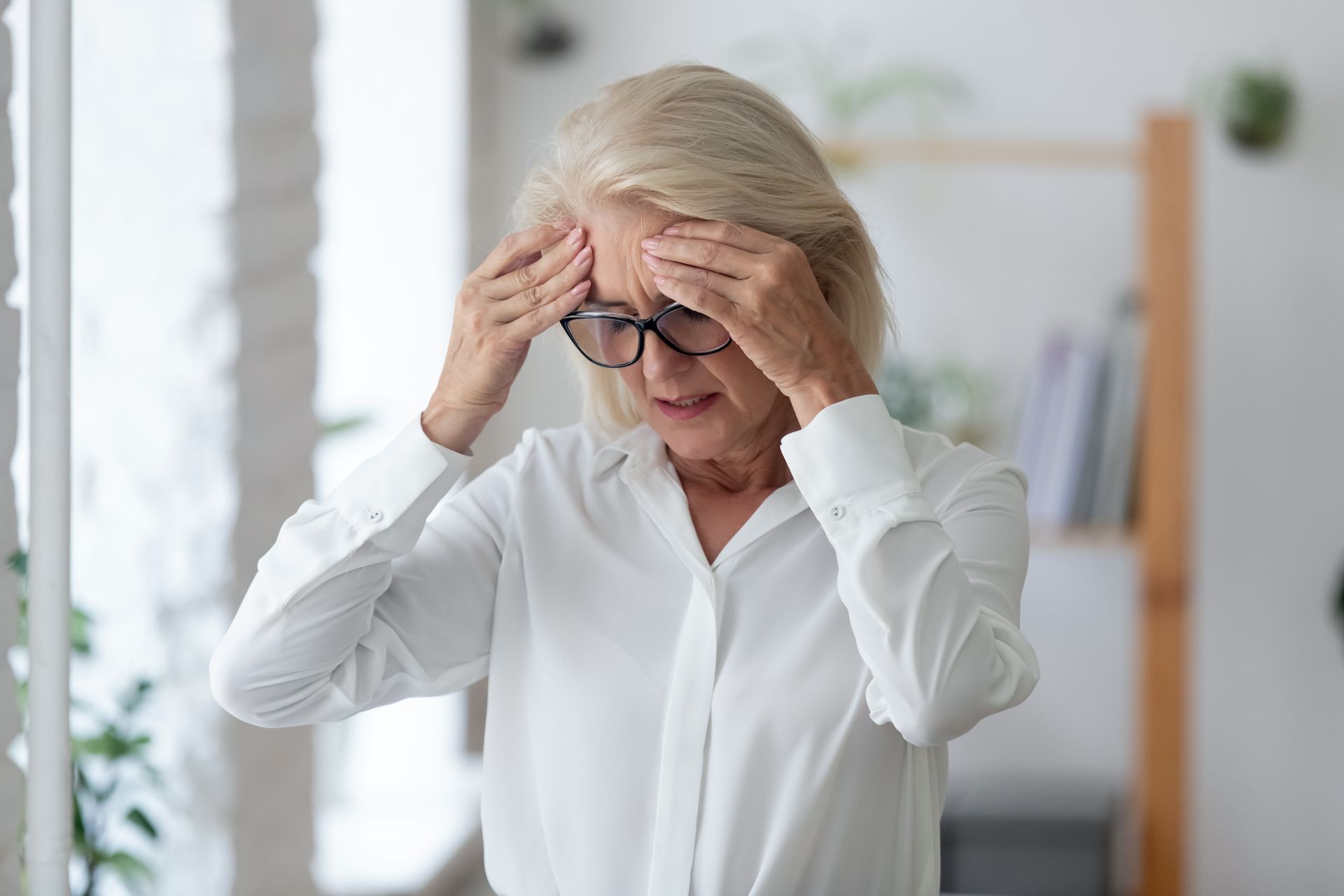 An elderly woman with glasses is holding her head in pain.