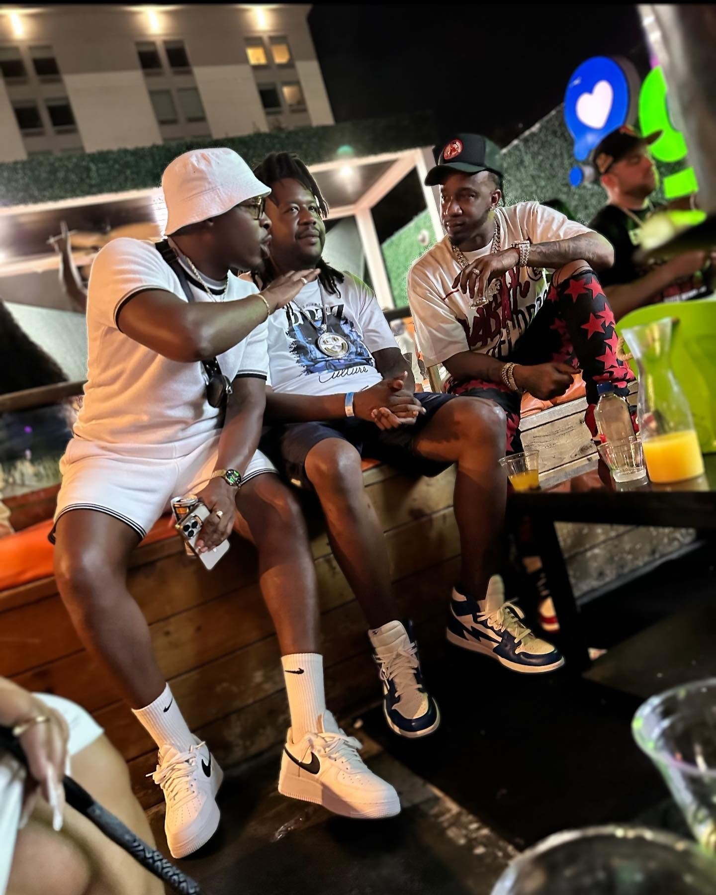 Three people seated outdoors, one pointing while conversing. Nighttime setting with lights and drinks visible.