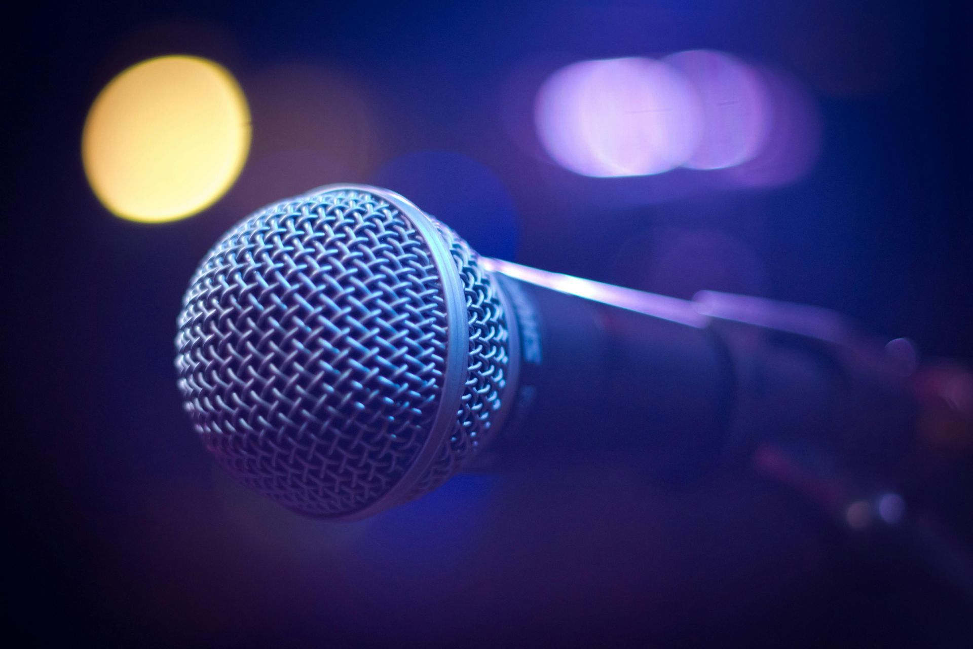 Close-up of a microphone on a stand, illuminated with blue and purple stage lights in the background.