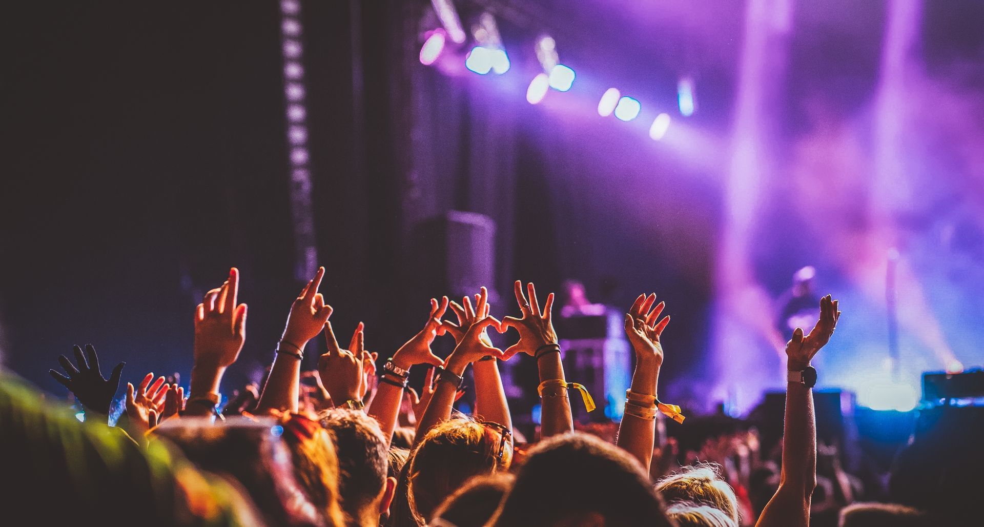 Concert crowd with raised hands, stage bathed in purple and white light.