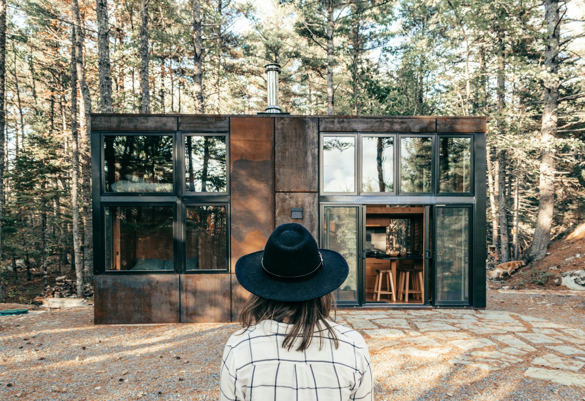 A woman in a hat is standing in front of a small house in the woods.