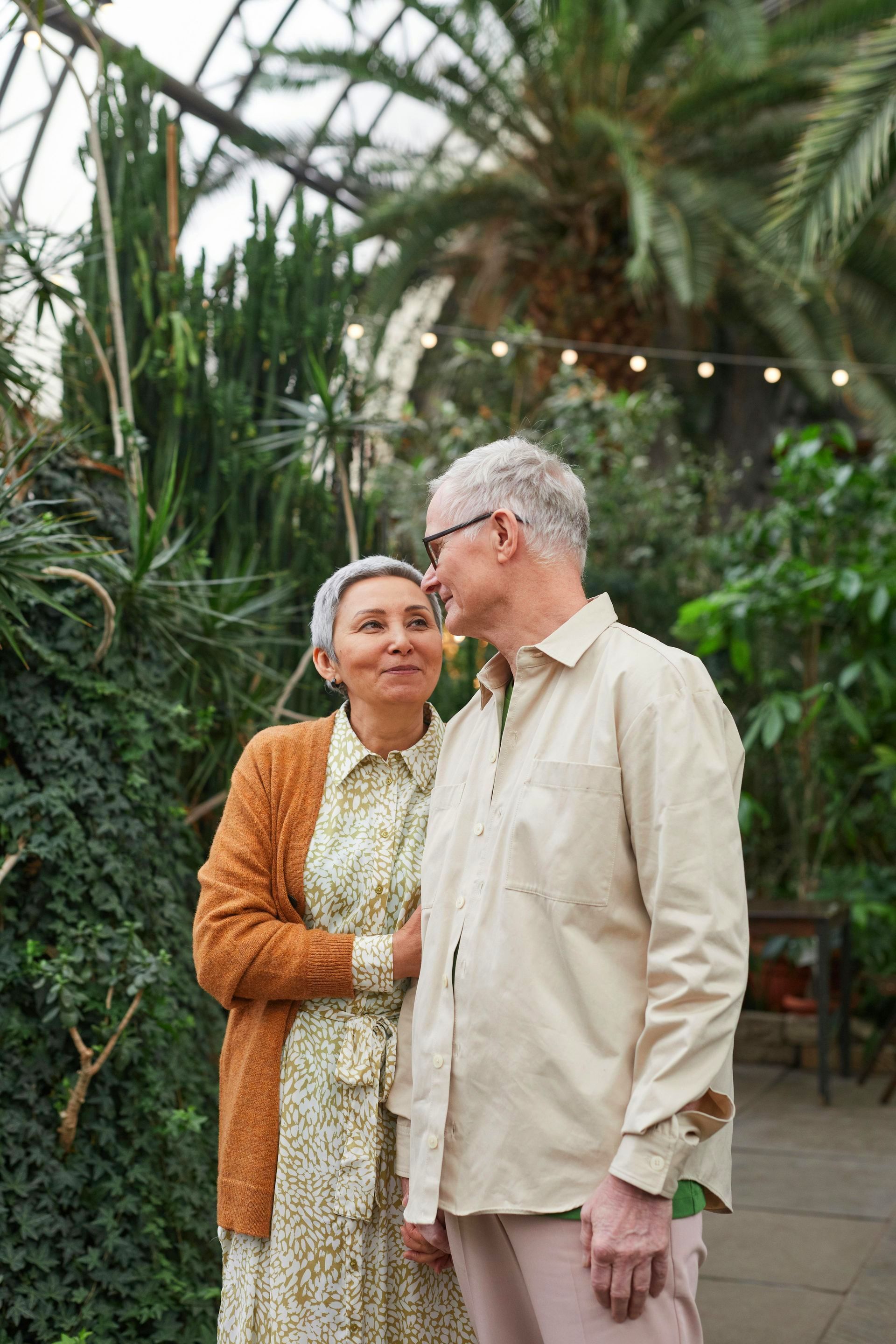 An elderly couple is standing next to each other in a greenhouse.