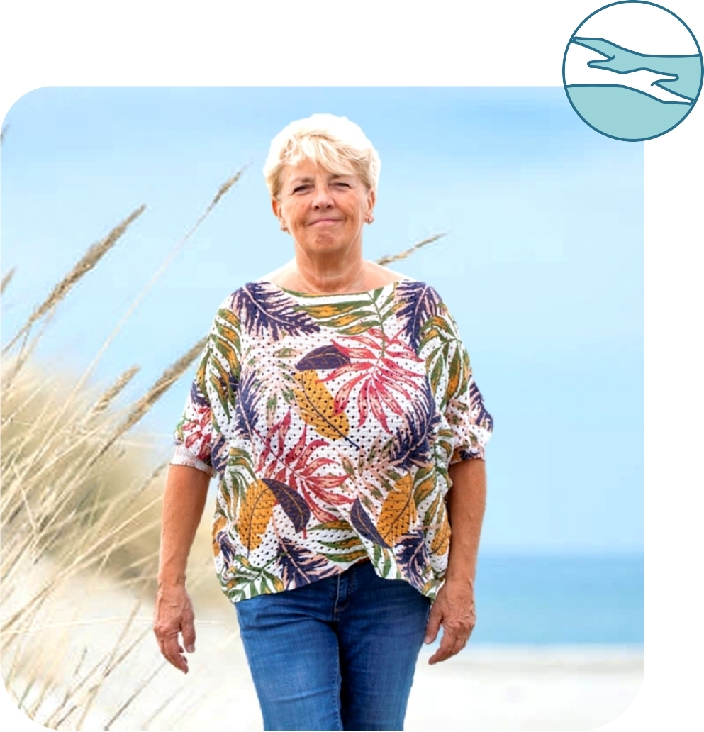 Een vrouw in een kleurrijk shirt en spijkerbroek loopt op een strand.