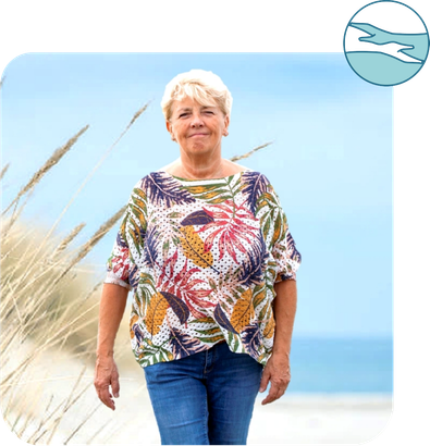 Een vrouw in een kleurrijk shirt en spijkerbroek loopt op een strand.