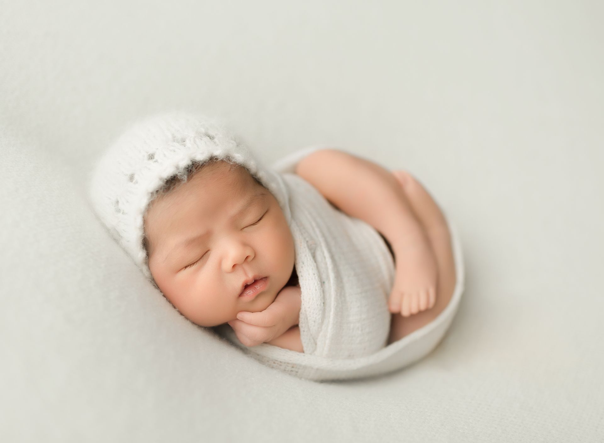 Newborn baby asleep in a white swaddle and hat, curled up on a white surface.