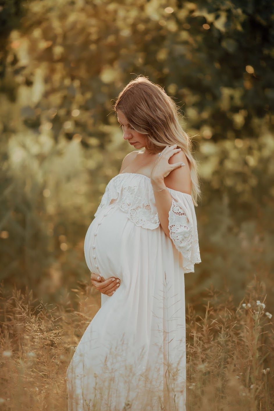 A pregnant woman in a white dress is standing in a field holding her belly.