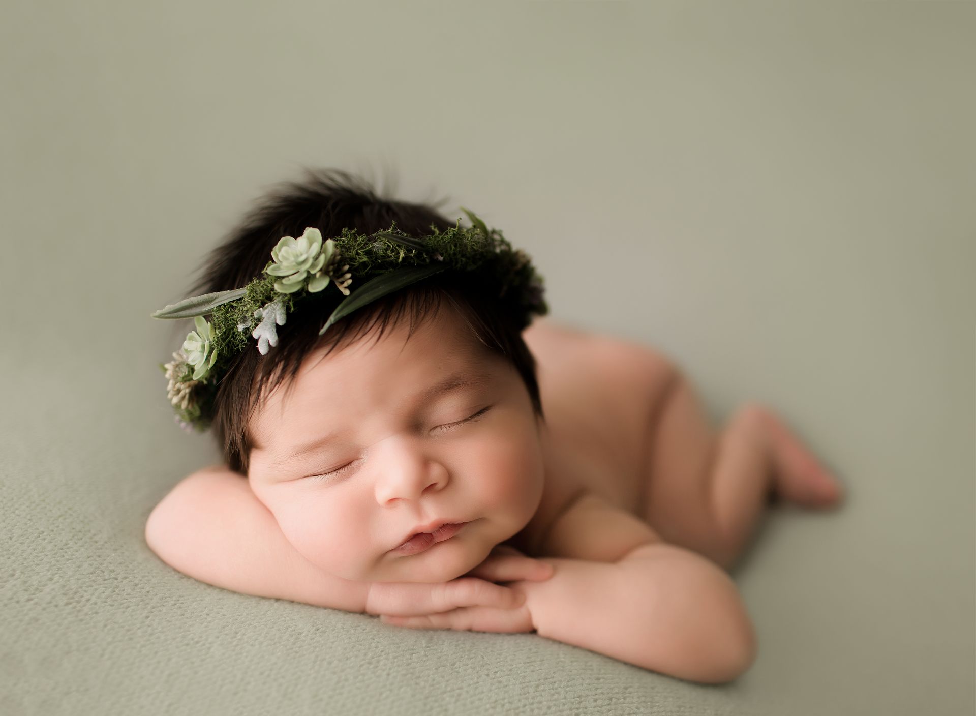 Newborn with dark hair and a leafy crown, lying on stomach with hands under chin, on a green surface.