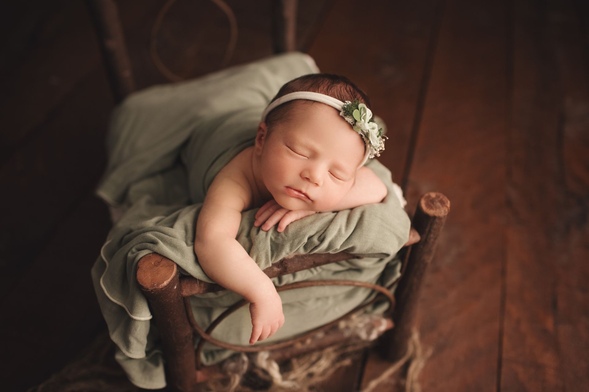 Newborn baby sleeping, draped in green fabric, resting on a rustic wooden chair, wearing a floral headband.