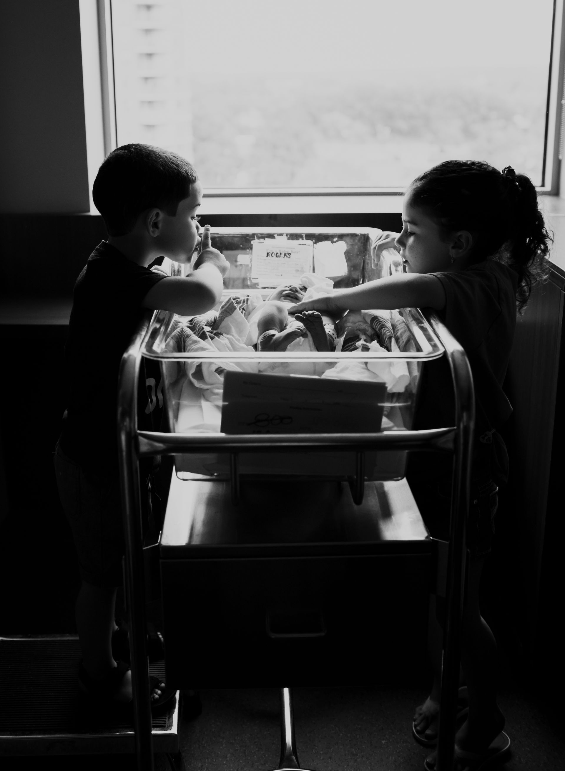 Two children looking into a hospital bassinet, lit by a window.