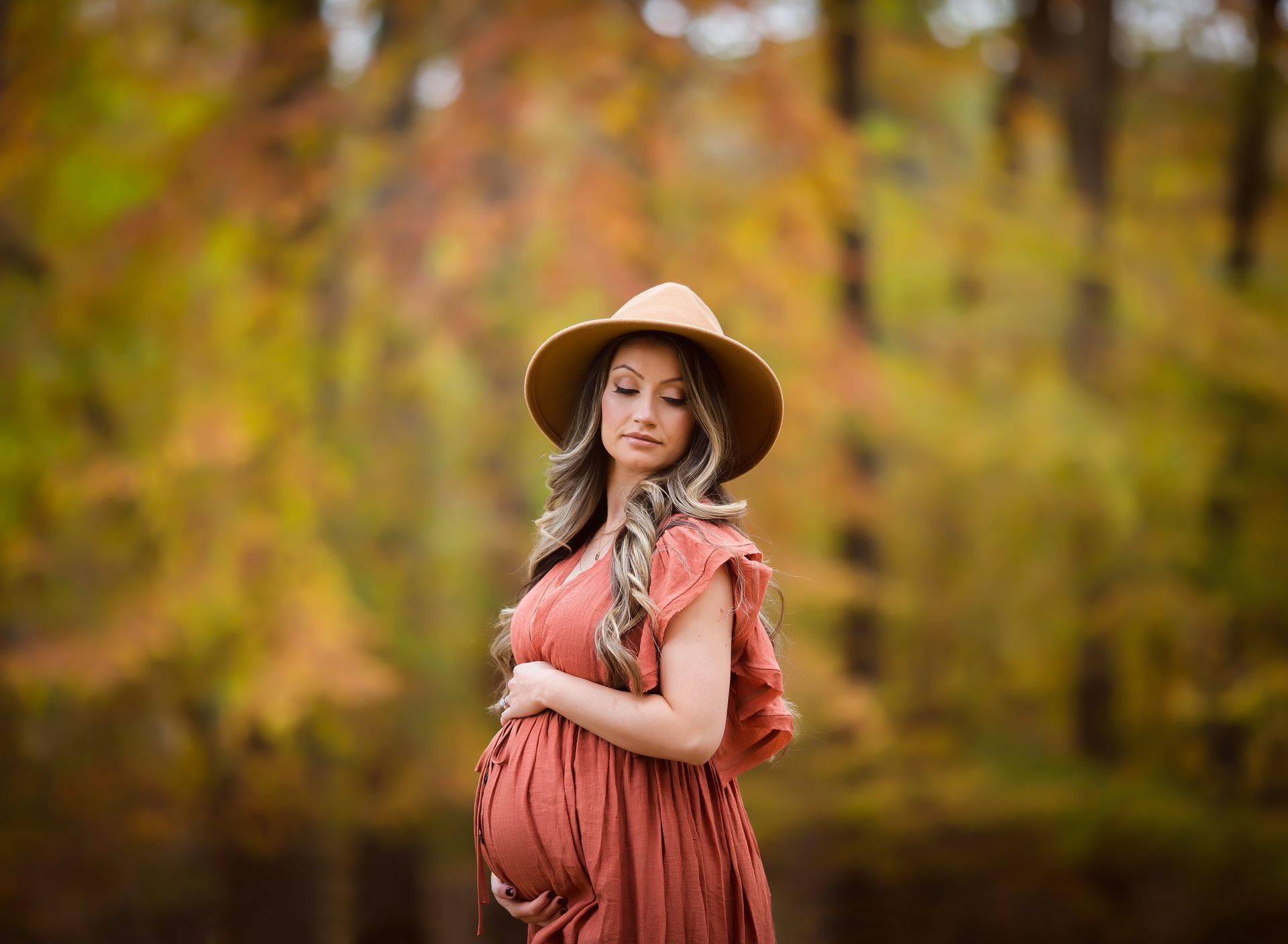 Pregnant woman in rust-colored dress and hat holds belly, standing outdoors with fall foliage.