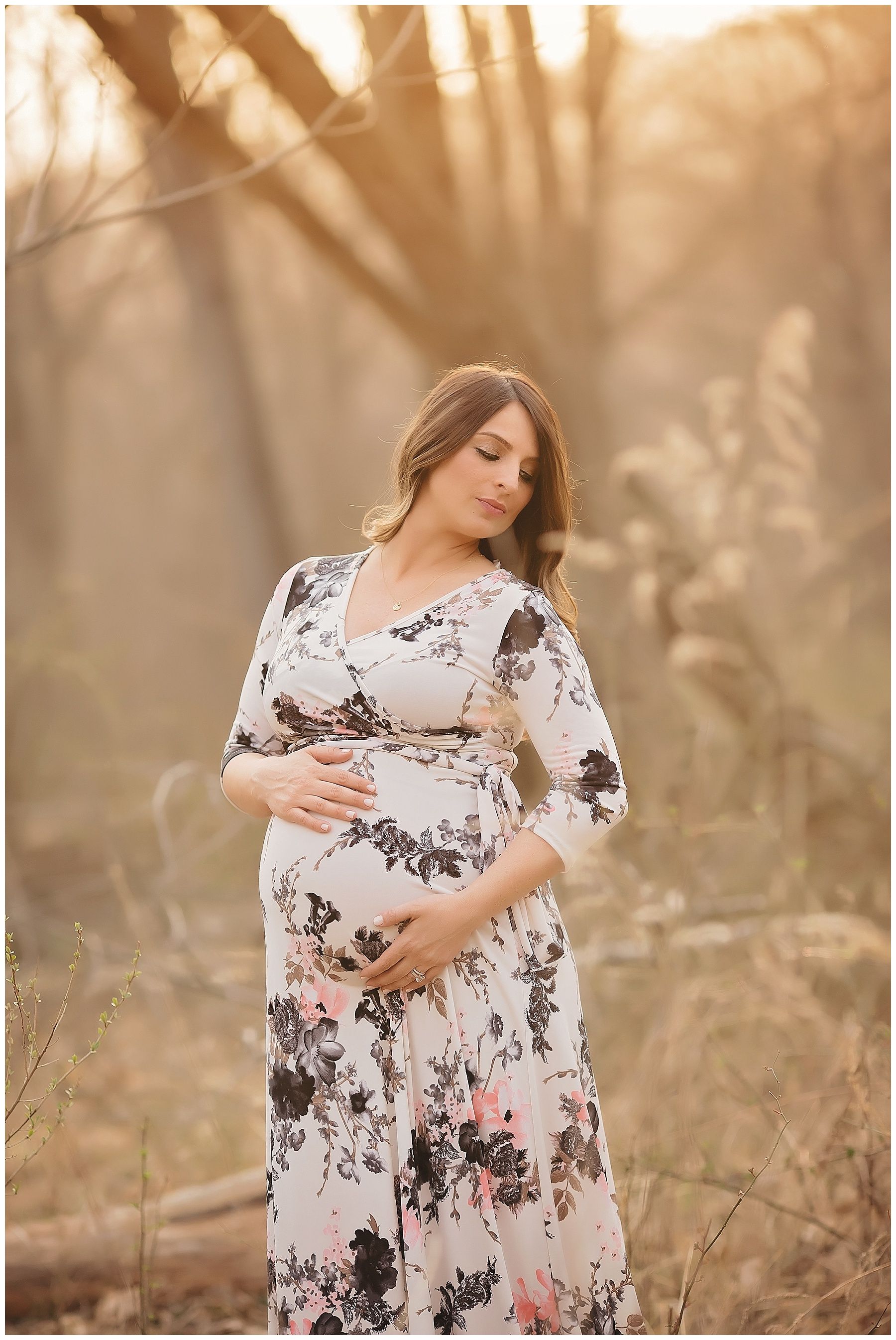 A pregnant woman in a floral dress is holding her belly in a field.