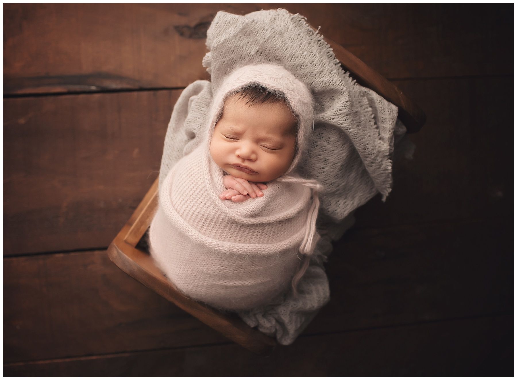 A newborn baby wrapped in a white blanket is sleeping in a wooden box.