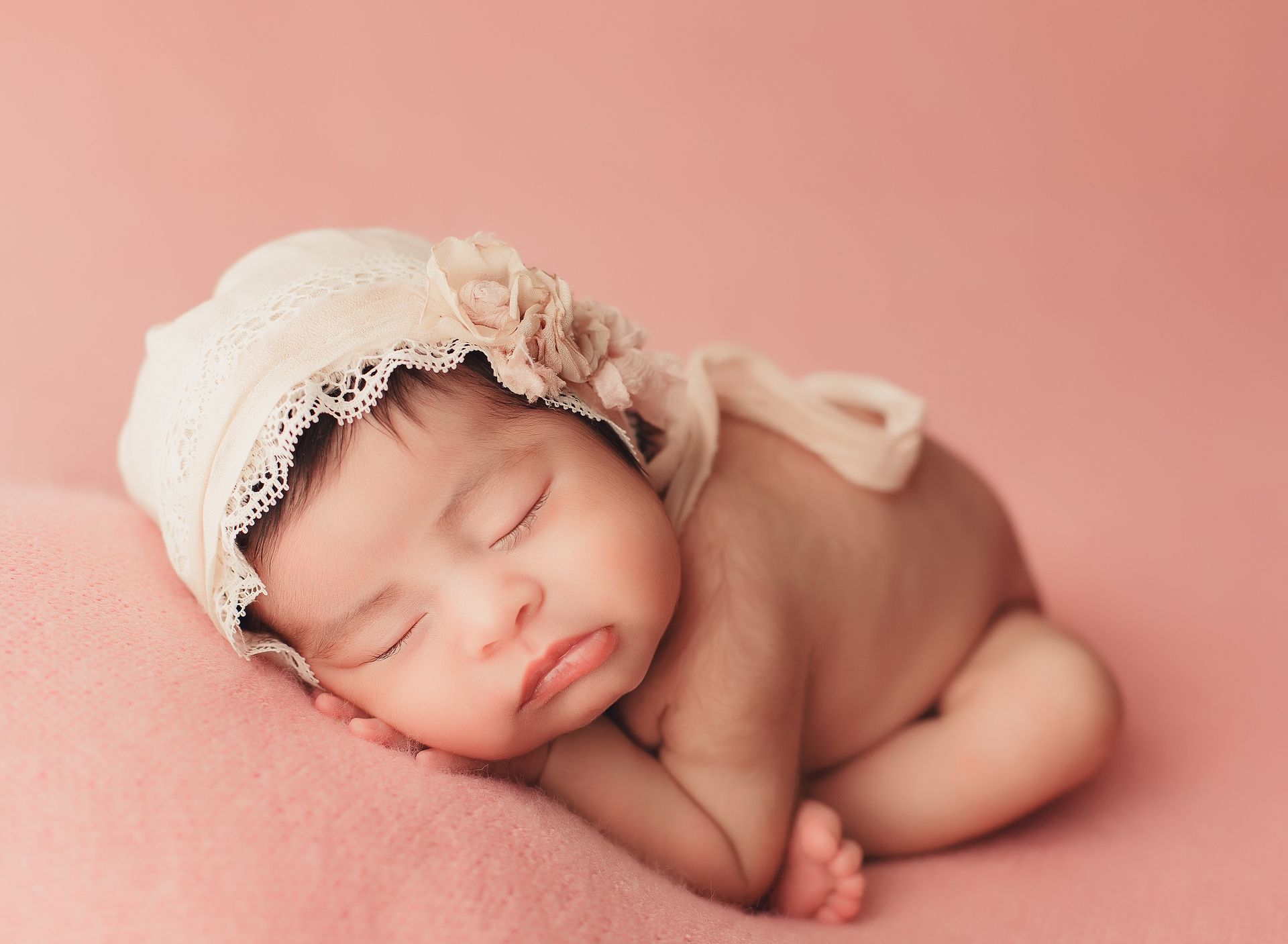 A newborn baby girl wearing a white hat is sleeping on a pink blanket.