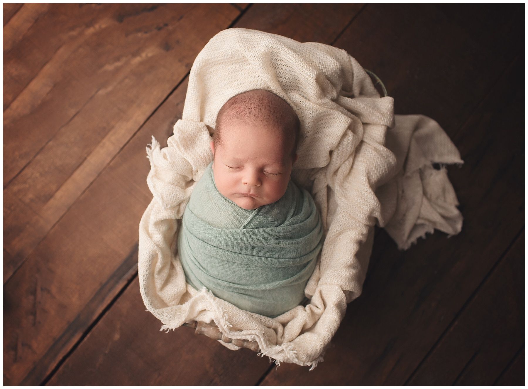 A newborn baby wrapped in a white blanket is sleeping on a wooden floor.