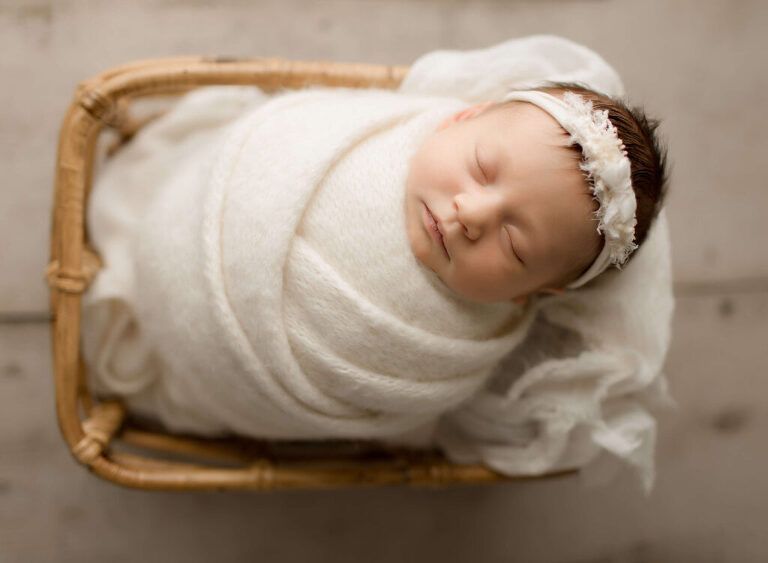 A newborn baby wrapped in a white blanket is sleeping in a wicker basket.