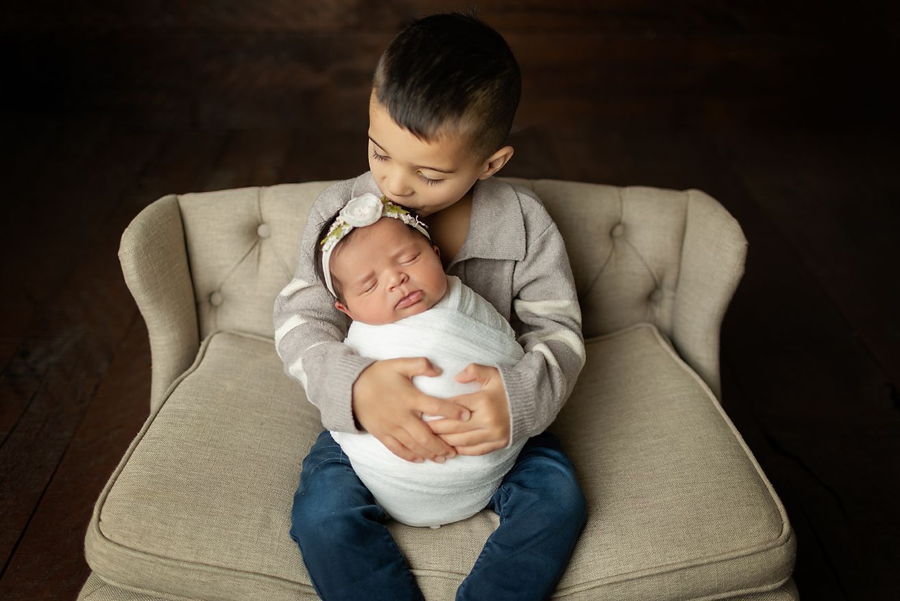 Older child holds a newborn, gently kissing its head. Both are seated on a vintage chair, dark wood background.