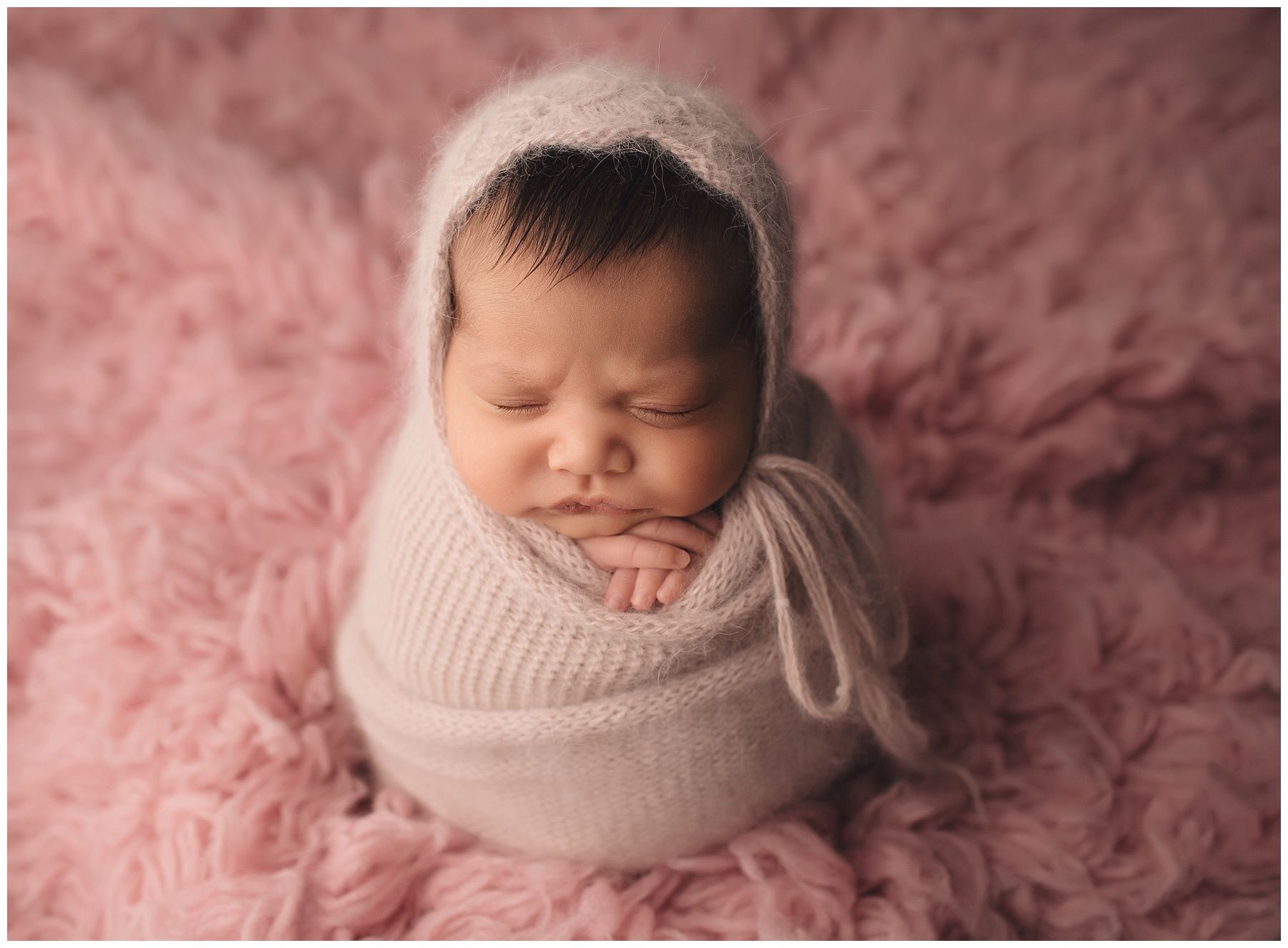 A newborn baby wrapped in a white blanket is sleeping on a pink blanket.