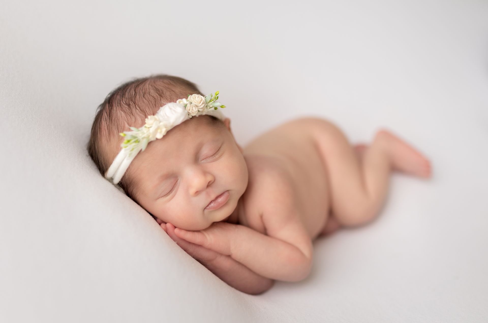 A newborn baby girl wearing a white headband is sleeping on a white blanket.