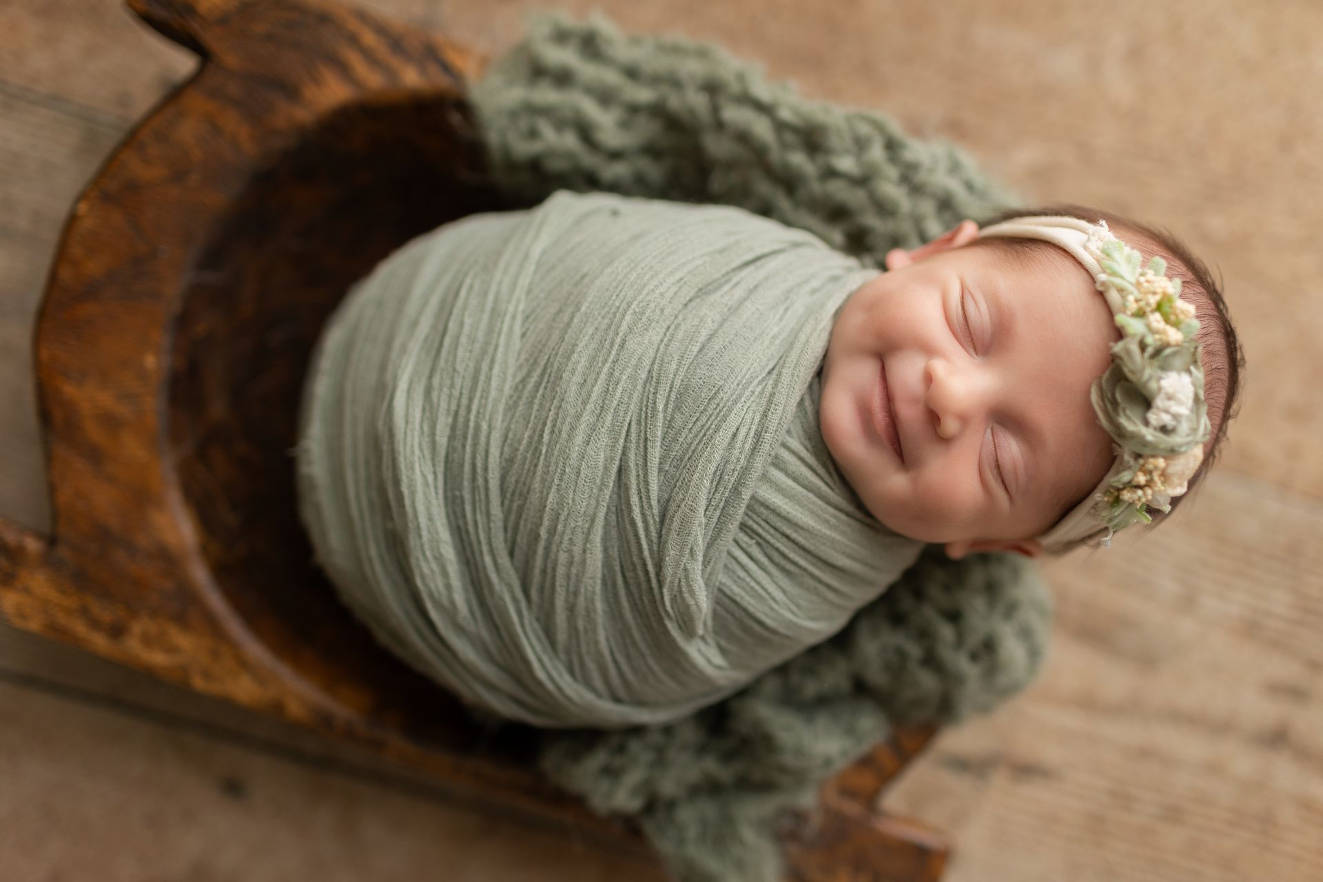 A newborn baby wrapped in a green blanket is sleeping in a wooden bowl.