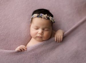 A newborn baby is wrapped in a purple blanket and wearing a flower headband.