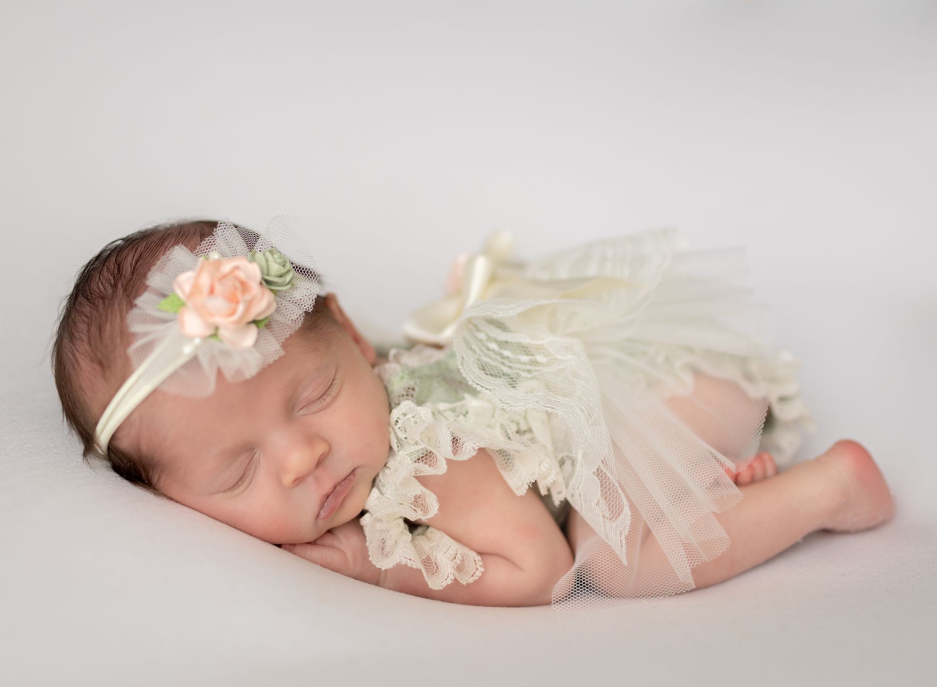 A newborn baby girl is sleeping on a white blanket wearing a lace dress and headband.