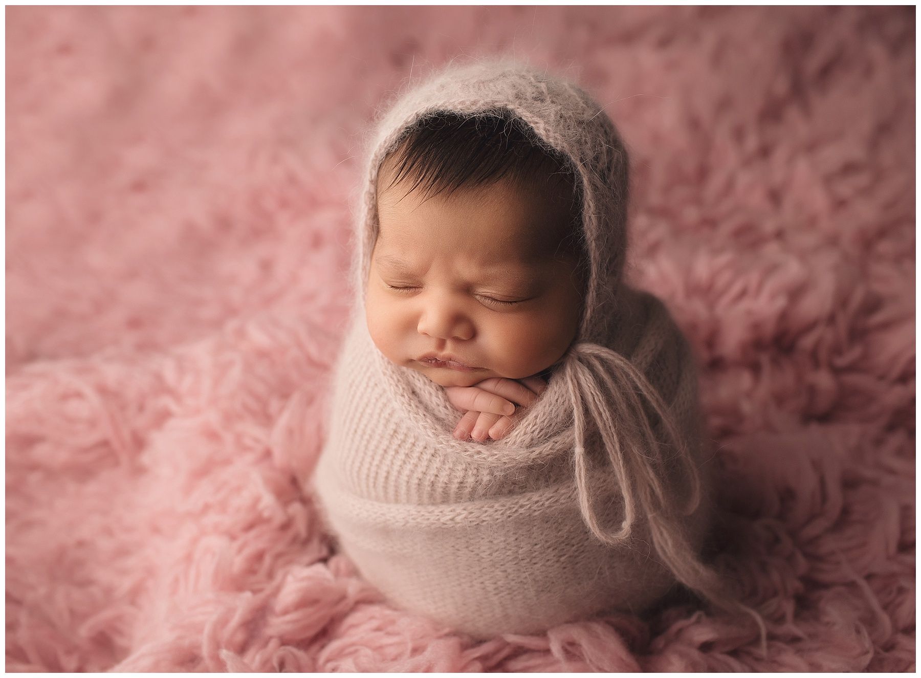 A newborn baby wrapped in a white blanket is sleeping on a pink blanket.