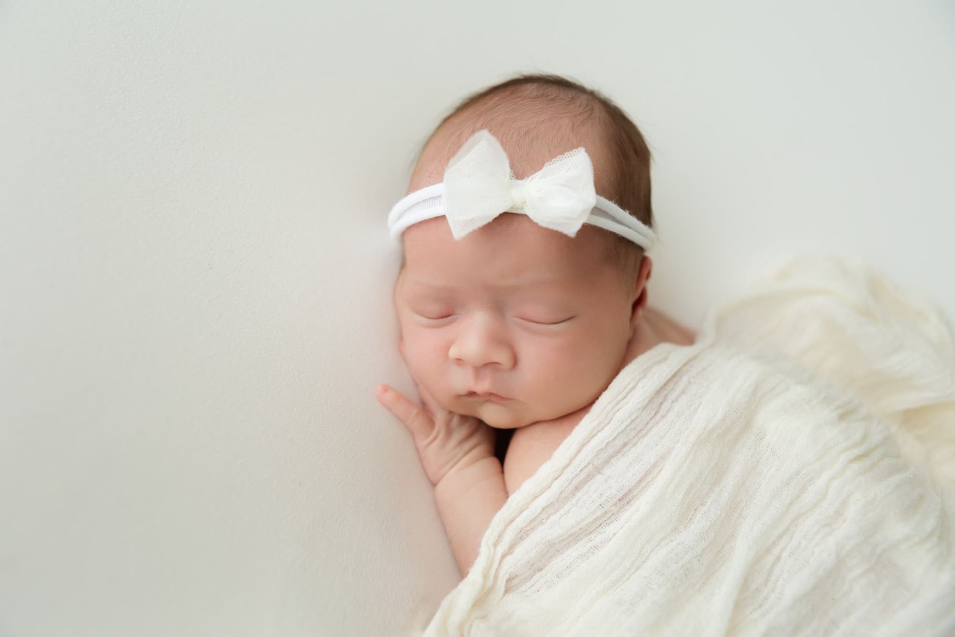Newborn baby sleeping, wearing a white bow headband, wrapped in a cream blanket.