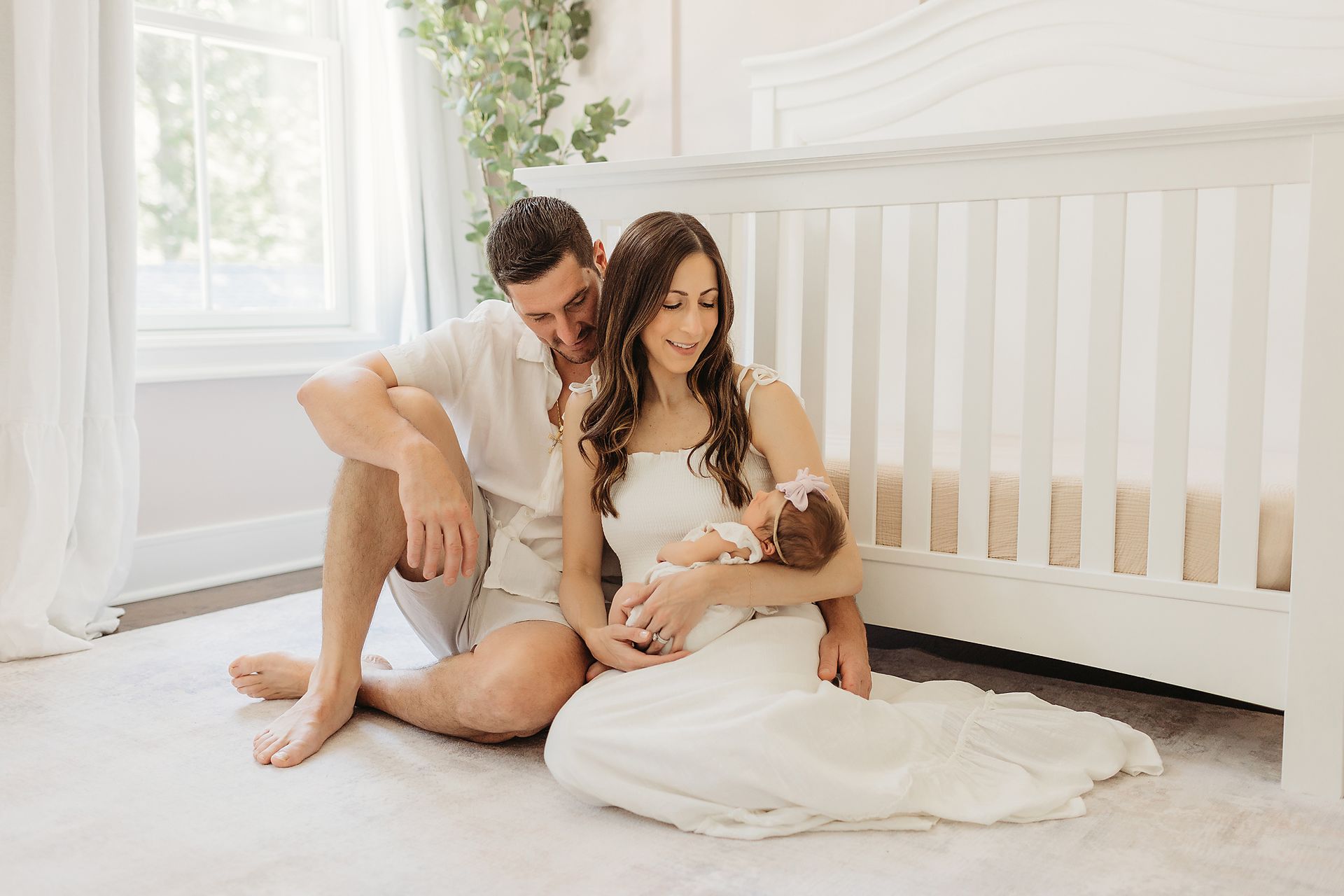 Parents sitting on floor, looking at newborn baby in white dress, near white crib, light room.
