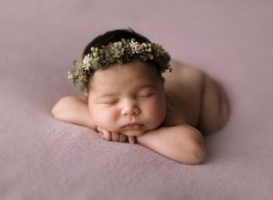 A newborn baby wearing a flower crown is sleeping on a pink blanket.