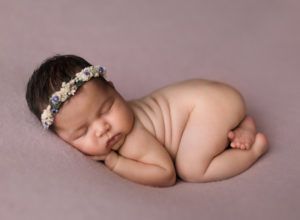A newborn baby wearing a flower headband is sleeping on a purple blanket.