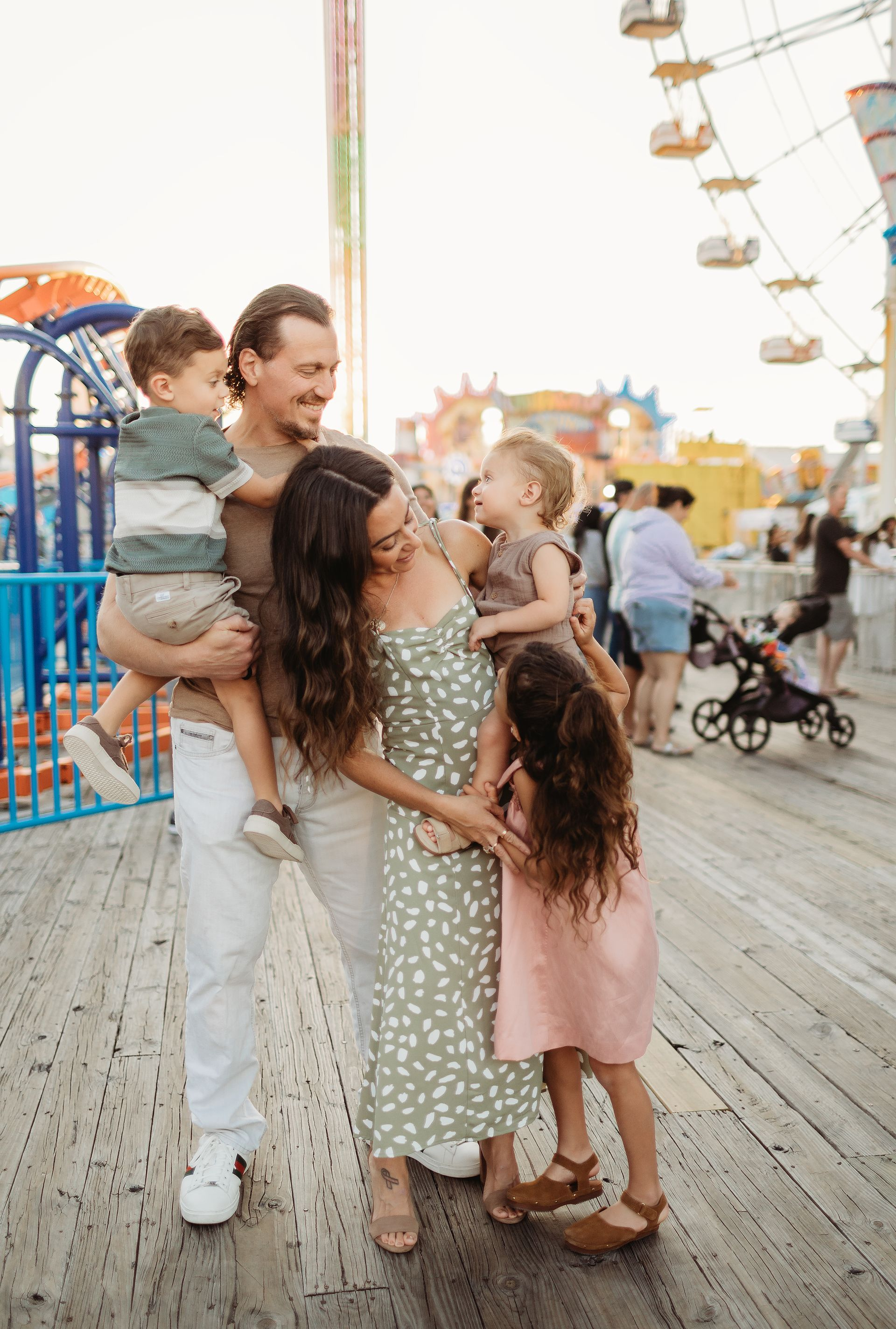 Family of five smiling at an amusement park. Father holding a child, mother embraces two children. Boardwalk and Ferris wheel in background.