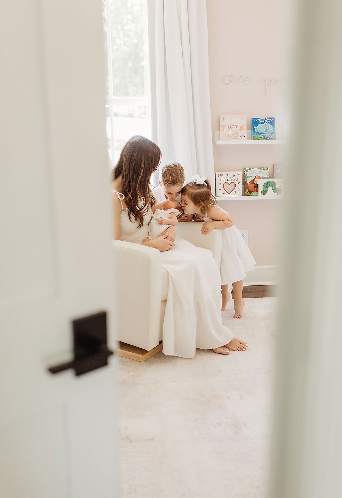 Woman holding a baby is surrounded by two children in a light-filled nursery.