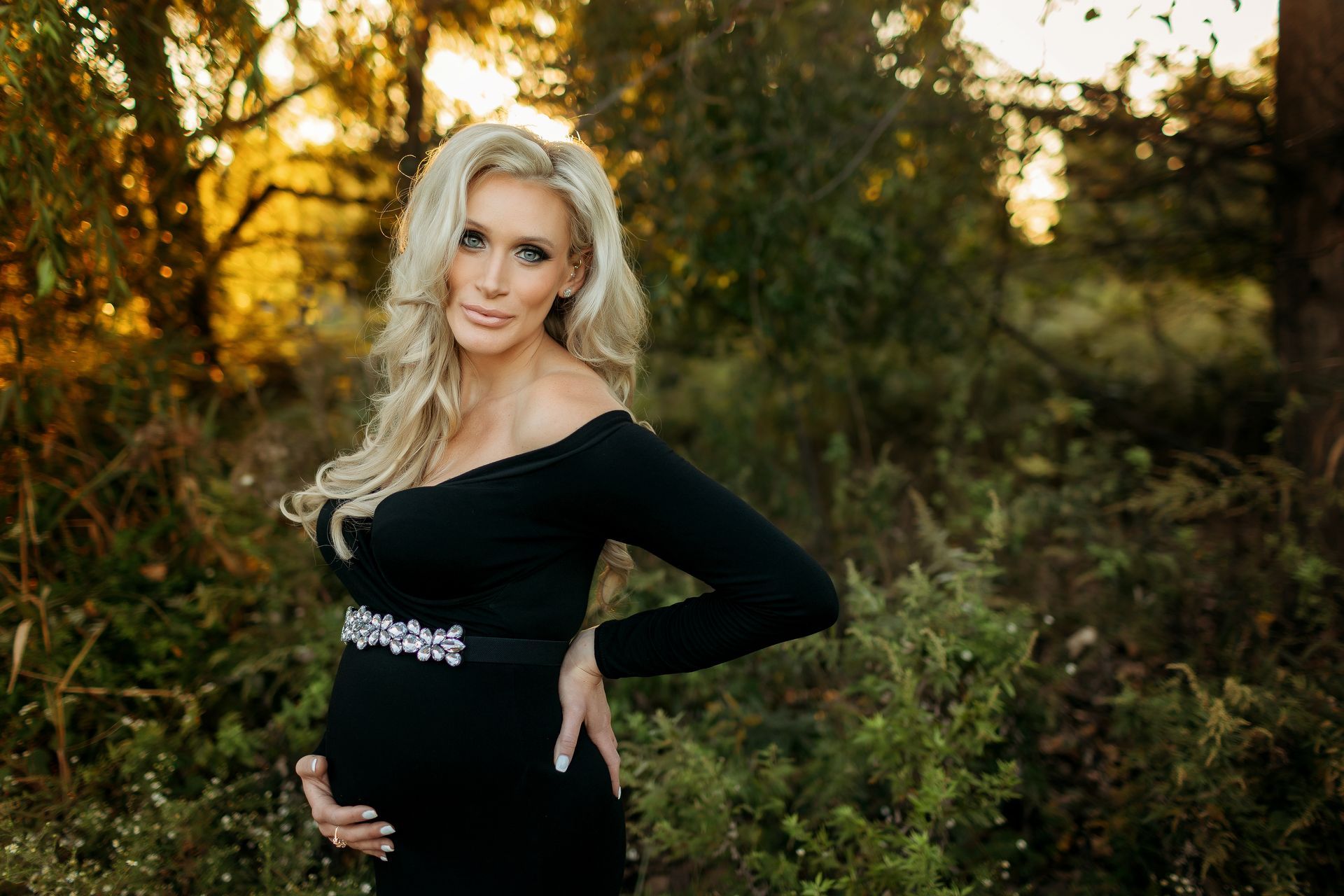 A pregnant woman in a black dress is standing in the woods holding her belly. Photo taken Near Leonia, NJ