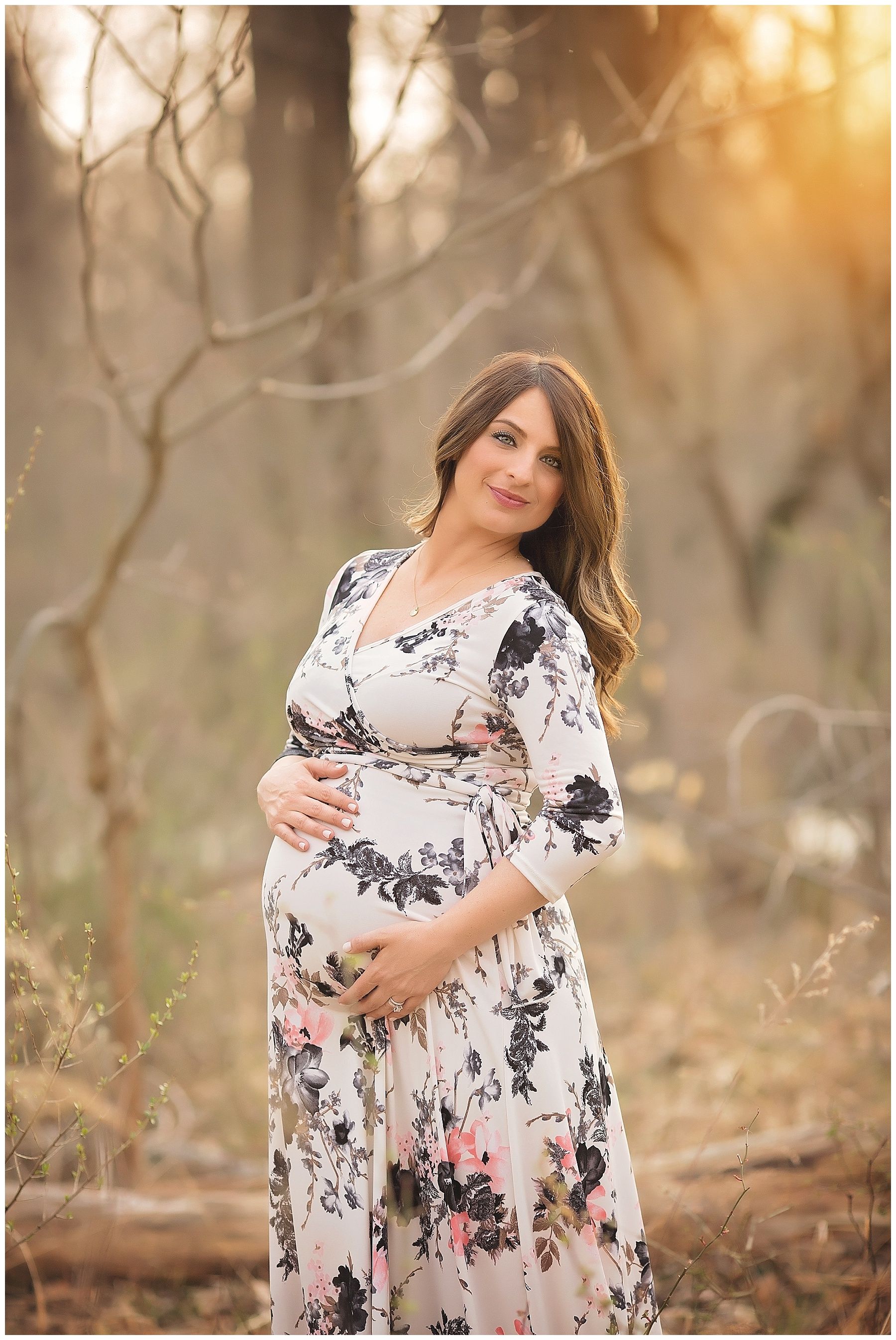 A pregnant woman in a floral dress is standing in the woods holding her belly.