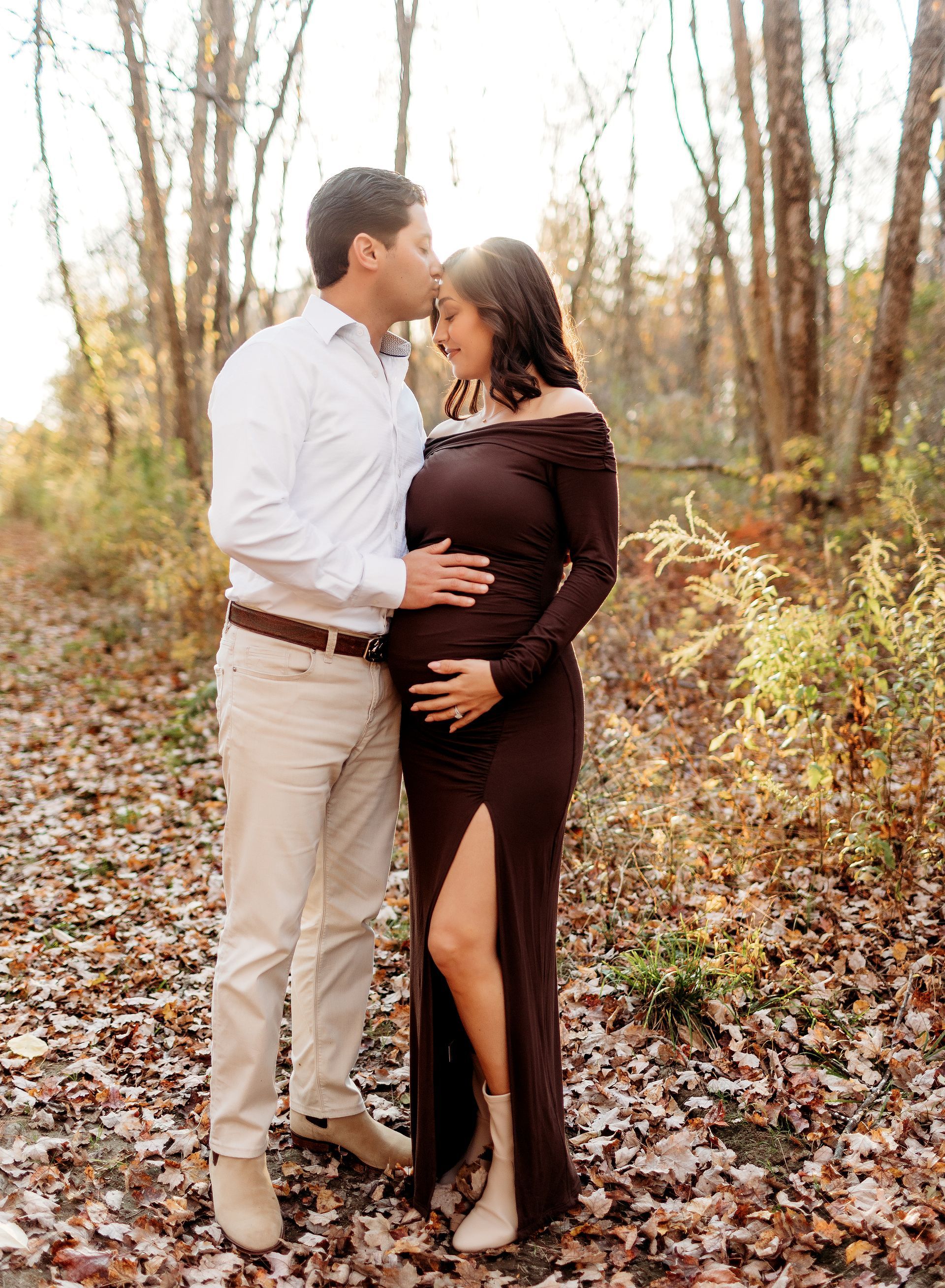 Couple in wooded area; man kisses pregnant woman's forehead, both hold her belly.