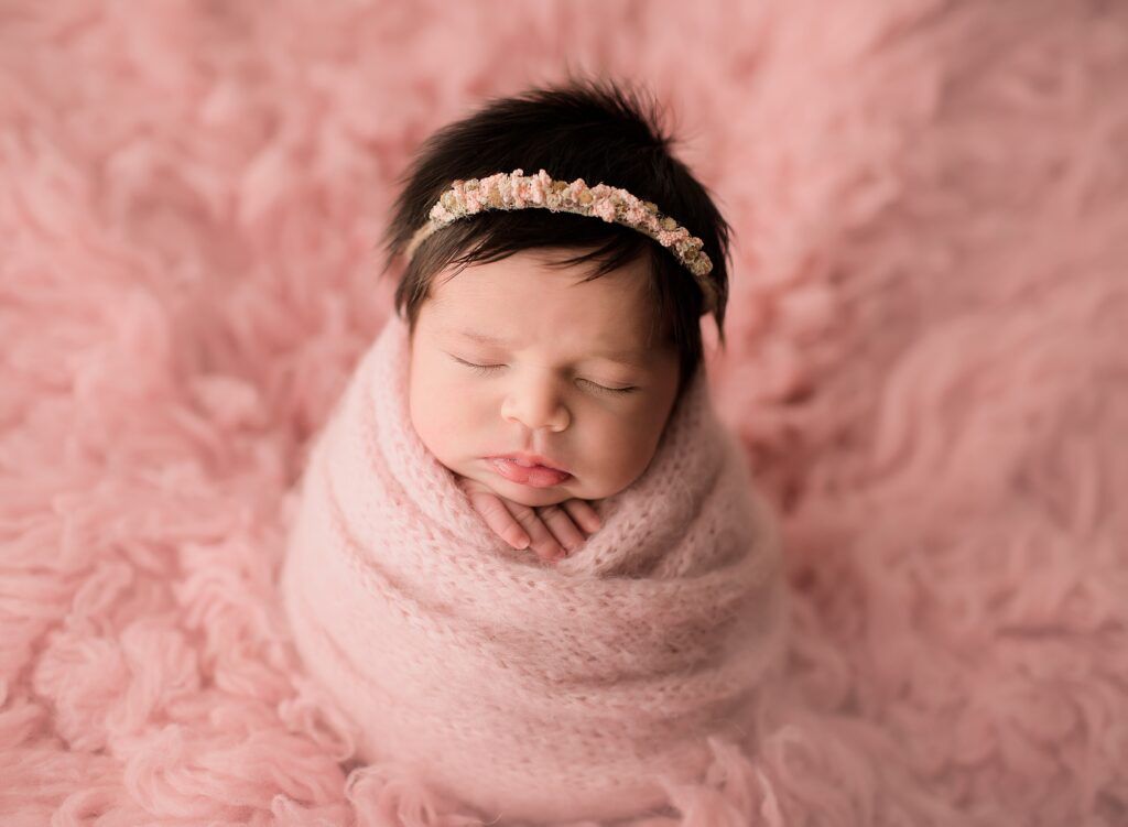A newborn baby girl is wrapped in a pink blanket and wearing a headband.