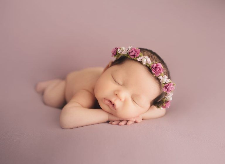 A newborn baby wearing a flower crown is sleeping on a pink blanket.